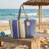 Een grote, handgemaakte gehaakte shopper met verticale strepen in verschillende tinten blauw, beige en wit, staand op een houten bankje op een zonnig strand.