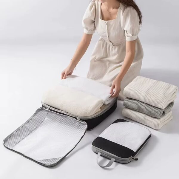 Een vrouw in een beige jurk plaatst gevouwen witte handdoeken in een gecombineerde mesh-en-stof compressiezak, met een stapel kleding ernaast.